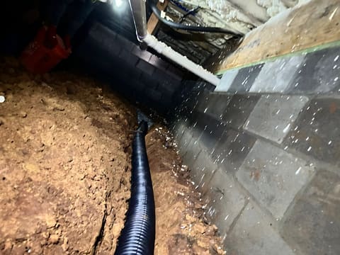 A new perforated drain line trenched into wet red clay along a crawl space foundation wall after rain